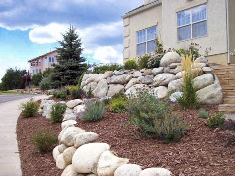 Multi-story residential building with landscaped front yard featuring large boulders, decorative rocks, and evergreen shrubs