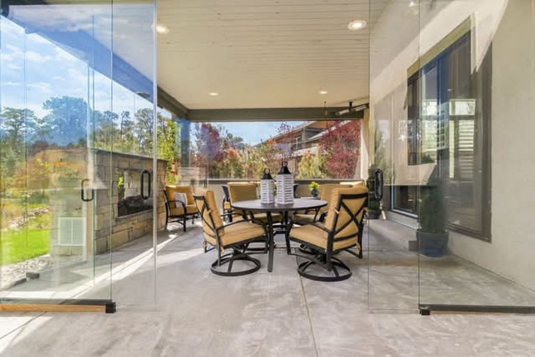 Spacious covered patio with dining table, chairs, ceiling fans, and views of autumn landscape through glass walls