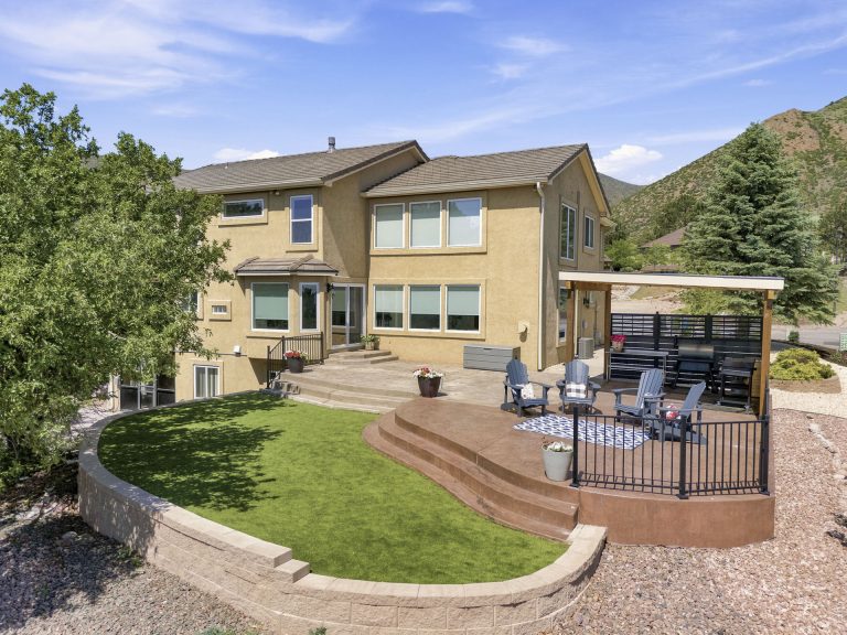 Modern yellow two-story house with manicured lawn, stone patio, and surrounding mountains