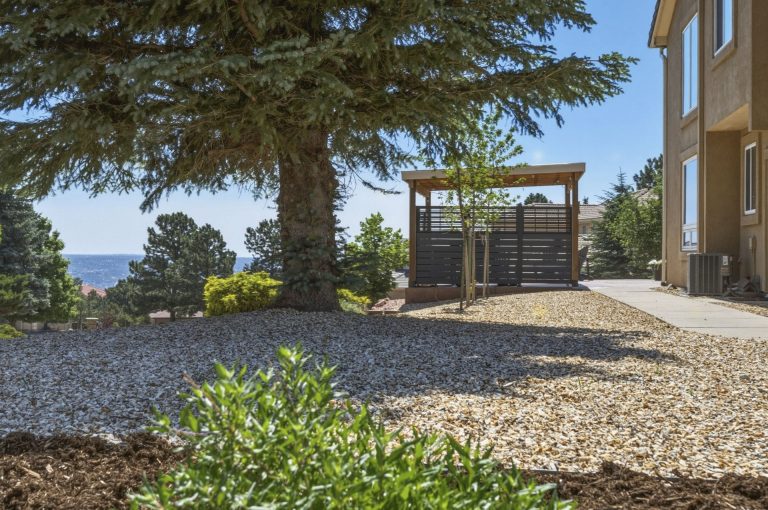 Wooden pergola and gazebo structures in a gravel patio area beside a large evergreen tree and house with water view