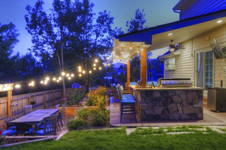 Backyard patio at dusk with string lights, stone seating area, modern house, and landscaped garden