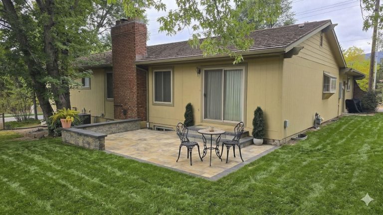 Tan single-story home with brick chimney, patio furniture, and manicured lawn surrounded by trees