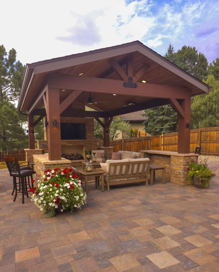 Covered wooden patio with seating area, potted flowers, and stone flooring surrounded by trees