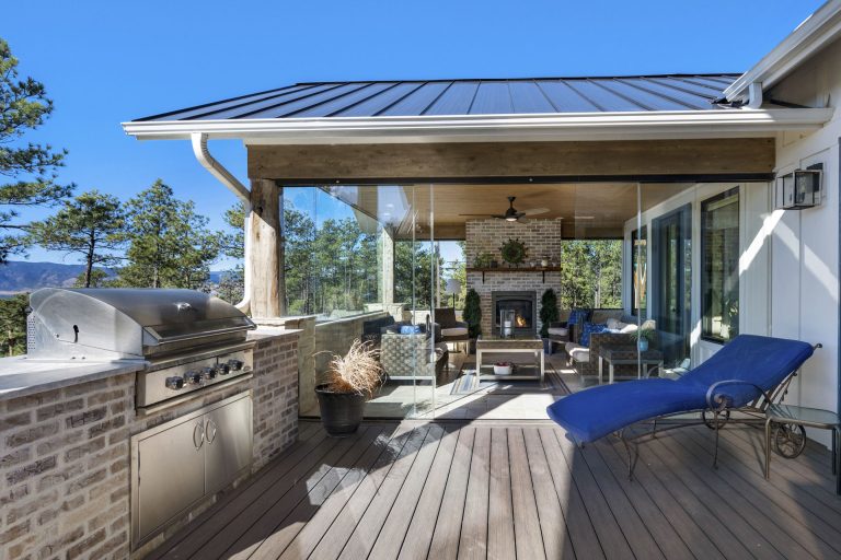 Modern covered deck with outdoor kitchen, grill, dining area, and lounge seating overlooking wooded landscape