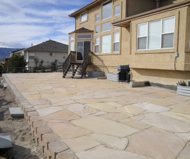 Newly installed stone patio with tan pavers in front of a two-story beige house with wooden deck stairs