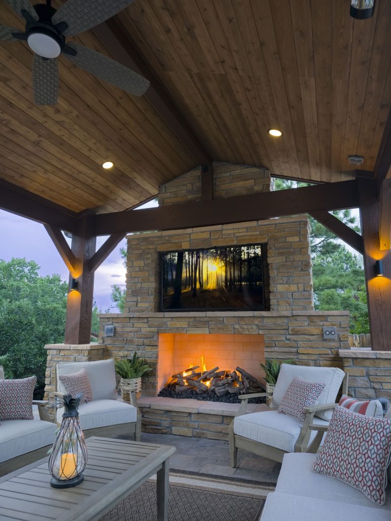 Covered outdoor patio with fireplace, seating area, wooden ceiling, and television mounted above brick fireplace