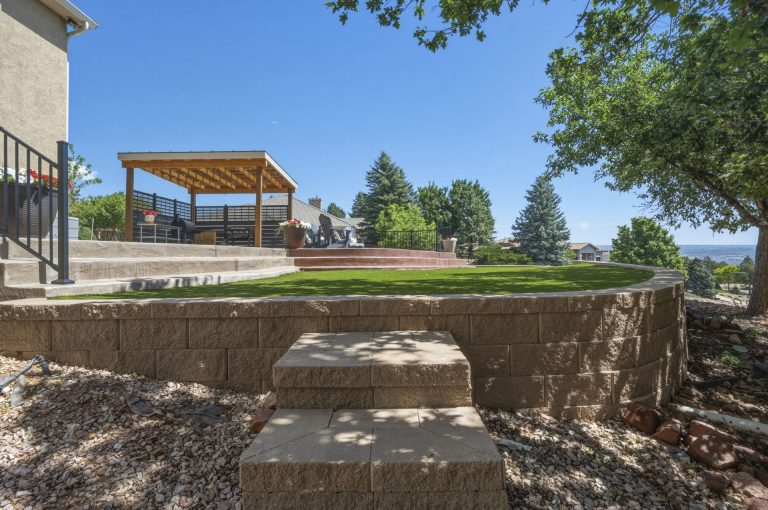 Modern backyard with stone retaining wall, wooden pergola structure, and manicured lawn area on sunny day