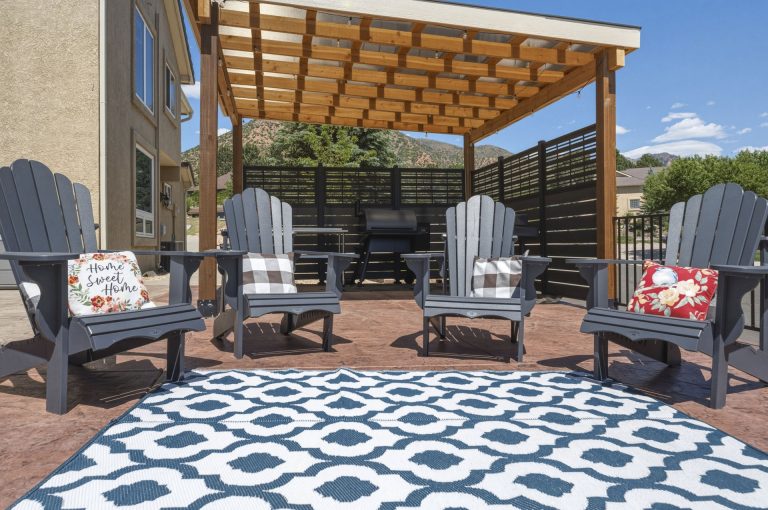Modern patio with wooden pergola, gray Adirondack chairs, geometric blue and white rug, and outdoor dining furniture