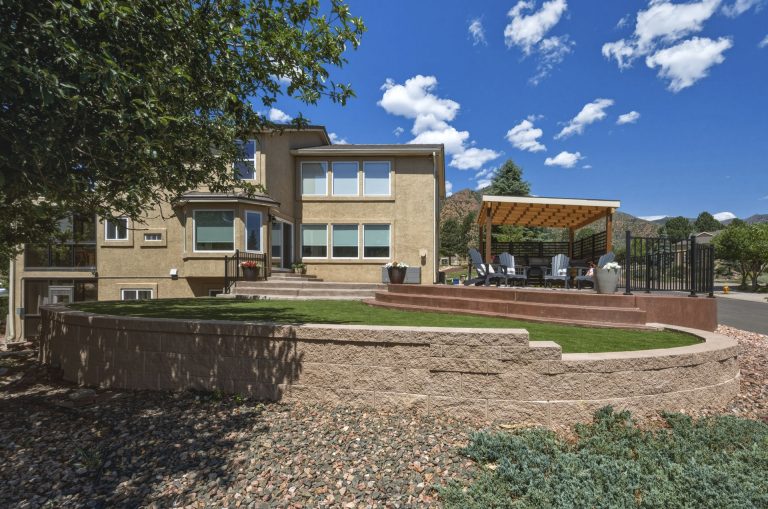 Modern residential home with tan brick facade, large windows, wooden pergola, and landscaped front yard with gravel and