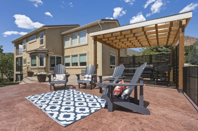 Modern home with wooden pergola, brick patio, outdoor furniture, and geometric patterned rug under blue sky