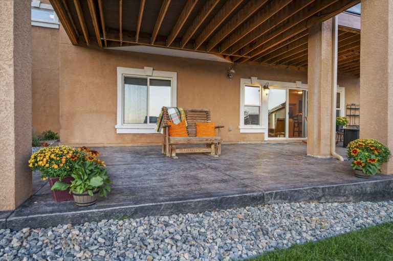 Covered patio with wooden beams, yellow Adirondack chair, potted flowers, and gravel landscaping