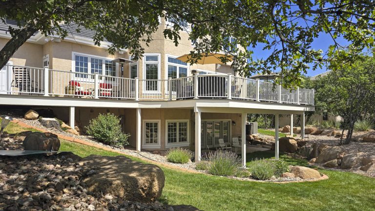 Two-story residential house with white railings and deck, stone foundation, surrounded by green lawn and mature trees