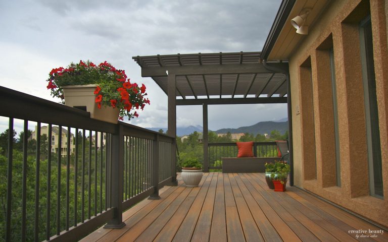 Wooden deck with black metal railing, pergola structure, and hanging flower baskets overlooking mountain landscape