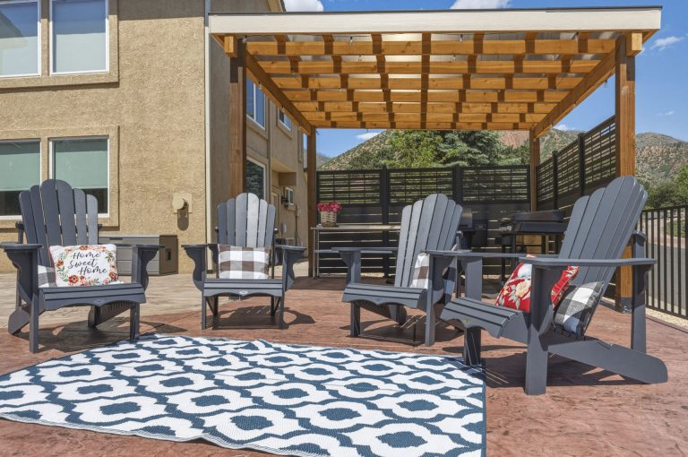 Covered patio with wooden pergola, Adirondack chairs, geometric patterned rug, and brick flooring