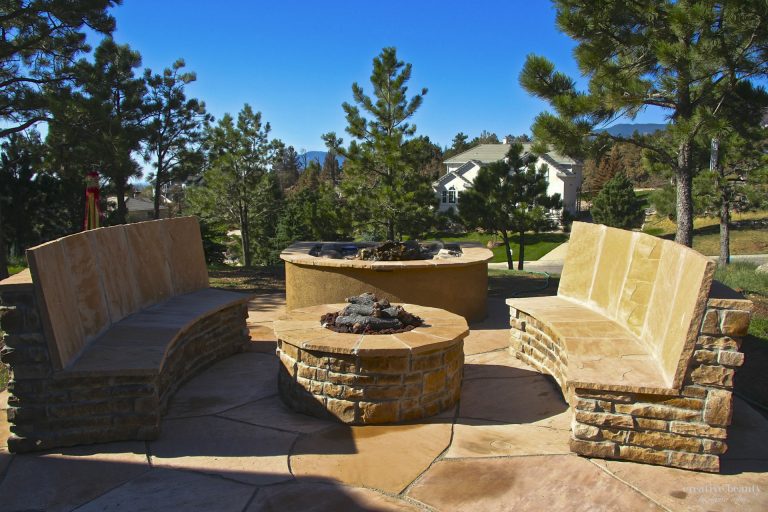 Circular outdoor fire pit seating area with stone benches and residential home visible in background among trees