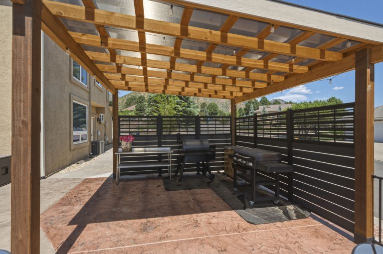 Wooden pergola with lattice roof and black privacy screen panels attached to tan brick house with patio area