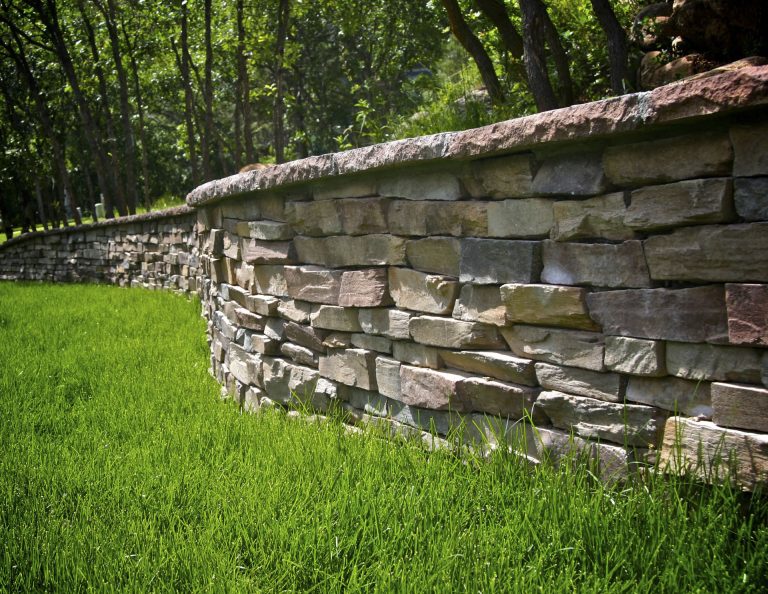 Rustic stone wall with horizontal layered slate blocks bordering a lush green lawn with forest trees in the background