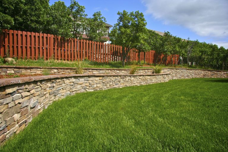 Manicured lawn with curved stone border and red wooden fence, backed by trees and blue sky