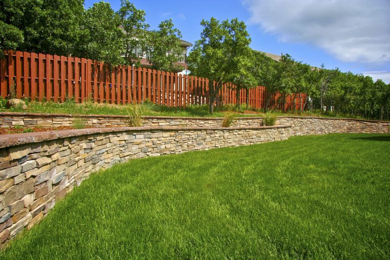Manicured lawn with curved stone border and red wooden fence backed by green trees under blue sky