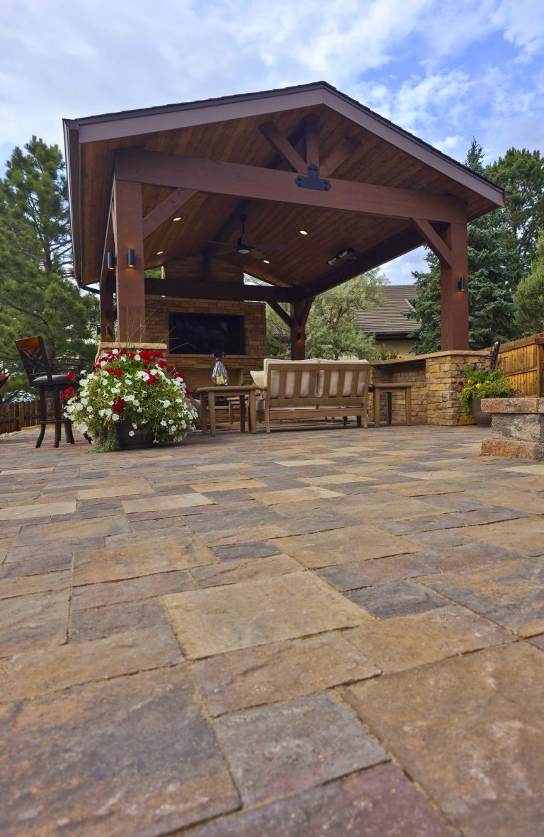 Wooden pavilion with ceiling fan and seating on a stone paver patio surrounded by landscaping