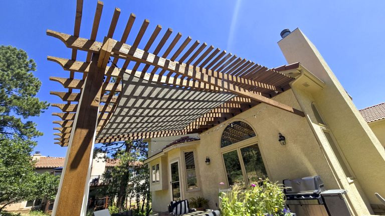 Wooden pergola attached to a beige residential house with landscaping and blue sky