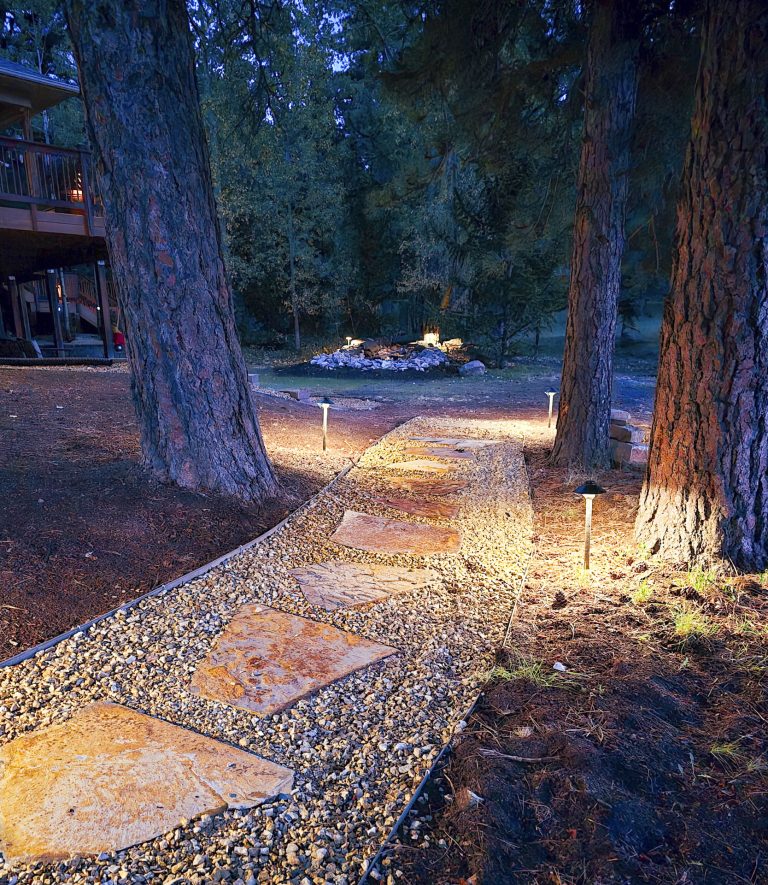 Illuminated gravel pathway winding through tall trees toward a lit outdoor seating area at dusk