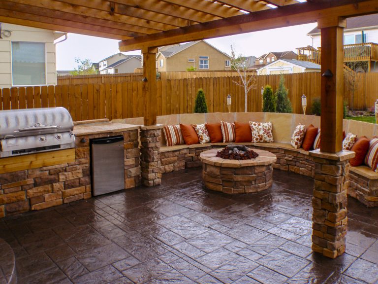 Covered patio with brick fire pit, outdoor seating, grill, and wooden pergola overlooking residential backyard