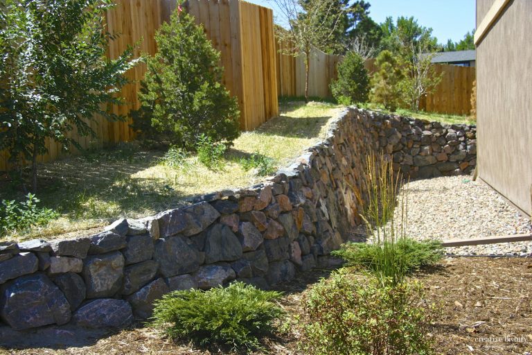 Backyard with wooden fence, stone retaining wall, gravel pathways, and landscaping shrubs under clear blue sky