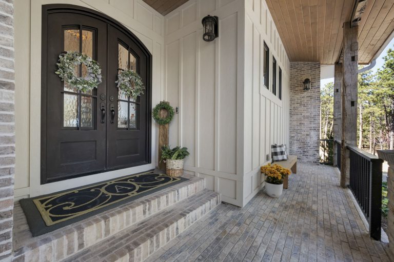 Front porch entrance with black double doors, white columns, brick pillars, and potted plants along a covered walkway