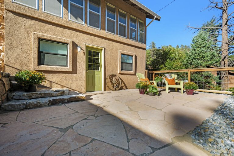 Beige stucco house with green door and flagstone patio surrounded by landscaping and trees