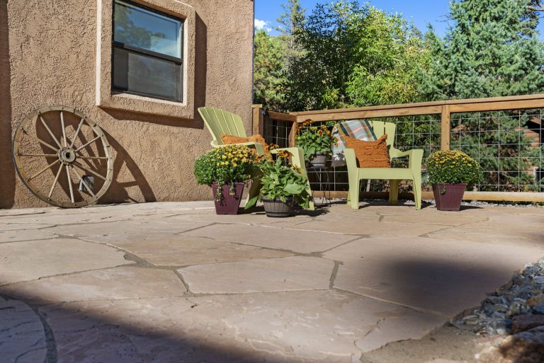 Rustic patio with concrete flooring, wooden wagon wheel, potted plants, and wooden bench against stucco building with window