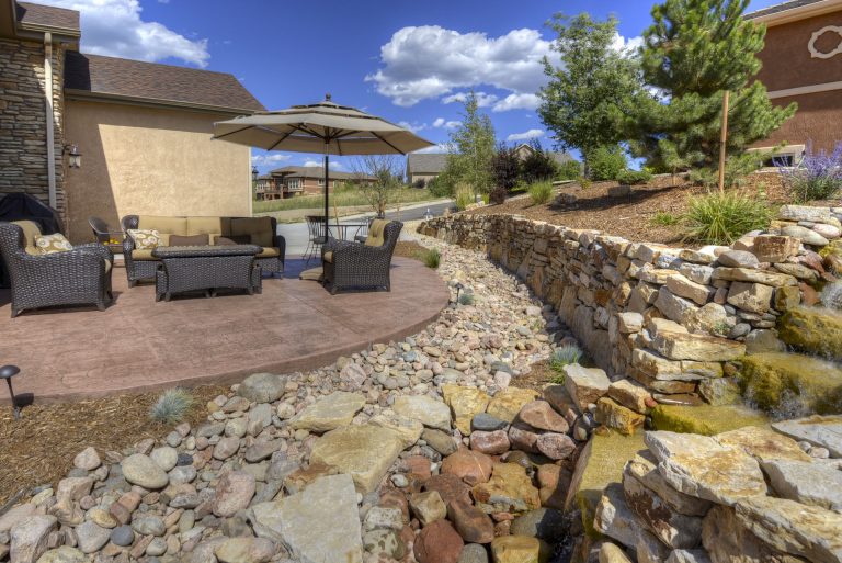 Modern backyard patio with stone fireplace, seating area, and landscaping along natural rock features