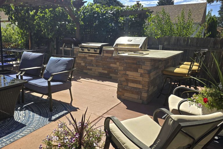 Outdoor patio with built-in brick grill, lounge chairs, and stone countertop surrounded by landscaping and trees