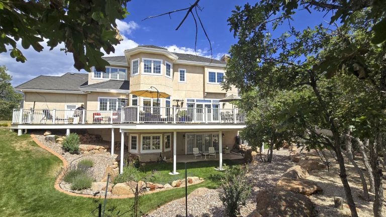 Yellow two-story house with wraparound deck surrounded by mature trees on sloped landscape