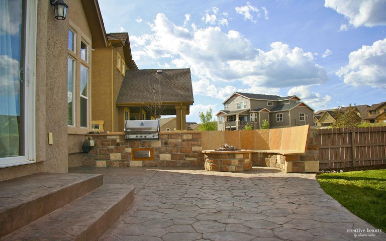 Spacious residential backyard with stone patio, wooden deck furniture, gazebo structure, and manicured lawn under blue sky