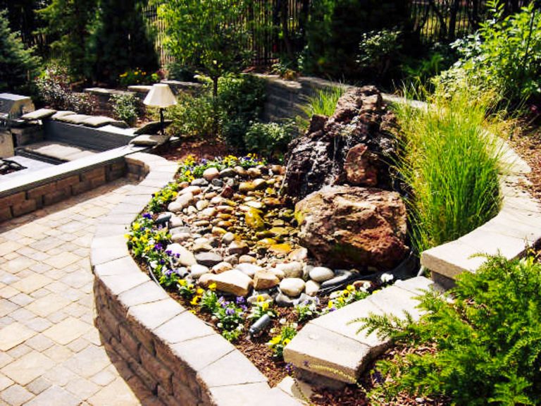 Raised stone garden bed with decorative rocks, small pond feature, and ornamental grasses bordered by pavers in a landscaped