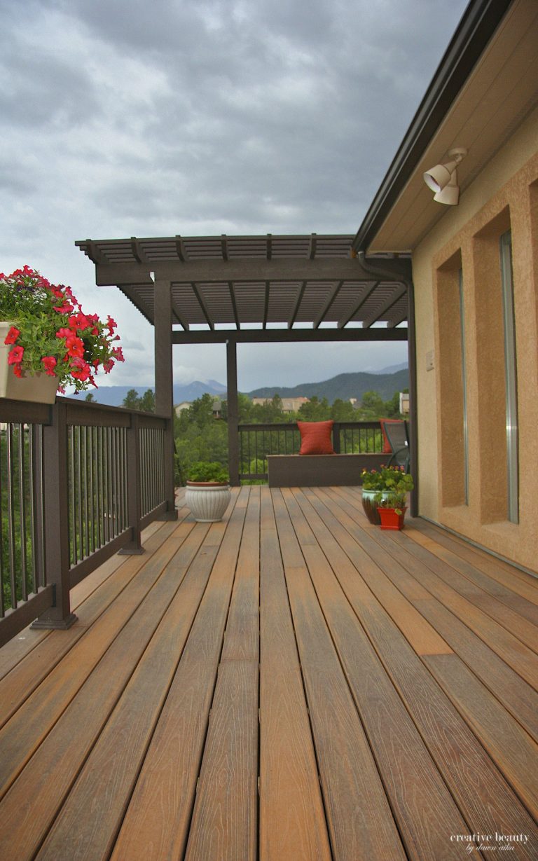 Wooden deck with pergola, potted flowers, and views of trees and mountains under cloudy sky