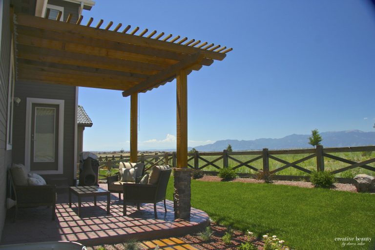 Wooden pergola attached to house with patio seating overlooking green lawn and distant mountains