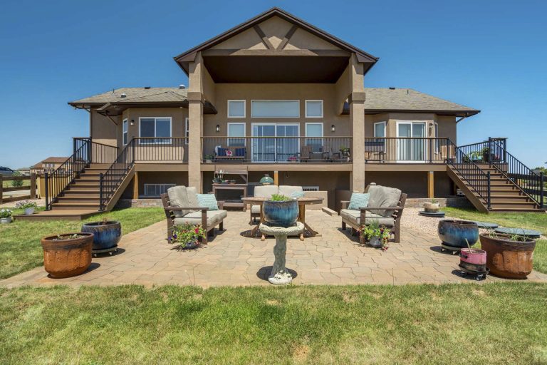 Large two-story tan house with covered patio, outdoor seating areas, and manicured lawn