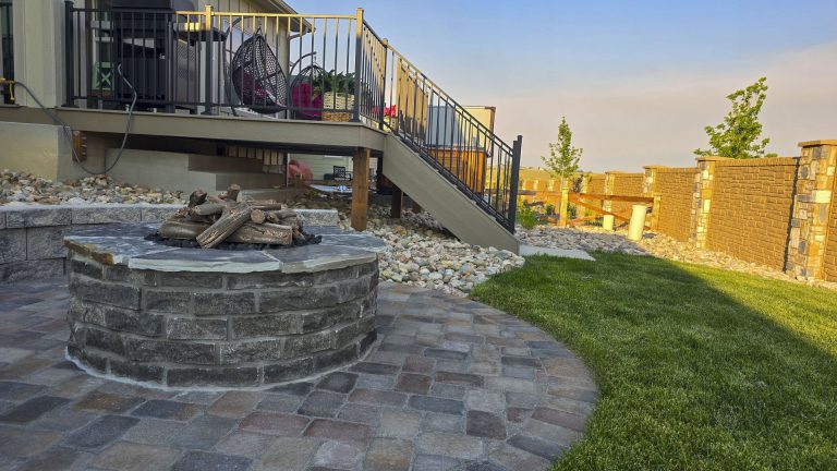 Backyard patio with circular stone fire pit, pavers, green lawn, wooden deck with railing, and stone wall fence at sunset