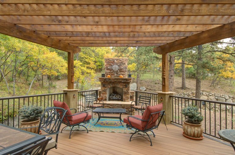 Covered deck with wooden pergola, fireplace, comfortable seating, and metal railings surrounded by trees