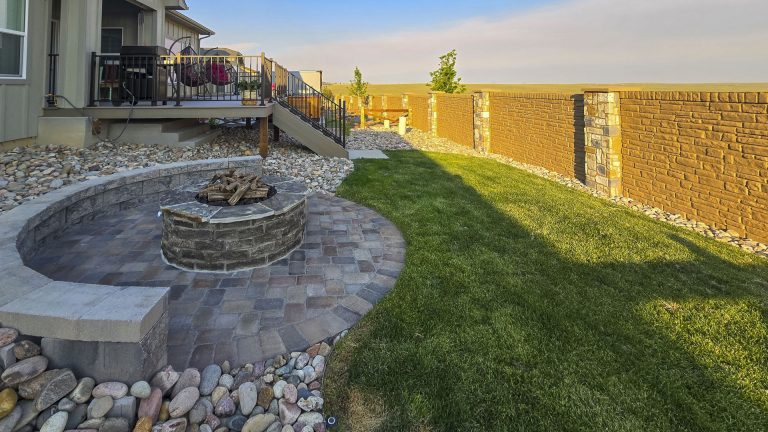 Backyard patio with circular fire pit, green lawn, decorative rock border, and wooden privacy fence on sunny day