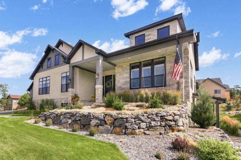 Modern two-story home with tan and dark exterior, stone foundation, landscaped yard with green lawn and decorative rocks