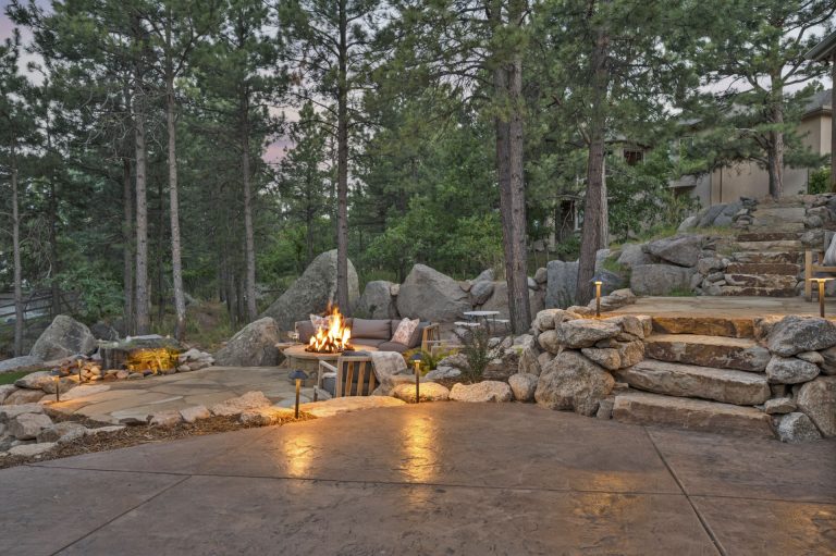 Outdoor fireplace with stone seating surrounded by logs and tall trees at dusk