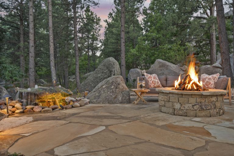 Outdoor patio with stone fireplace, fire pit, seating area, and surrounding pine trees at dusk