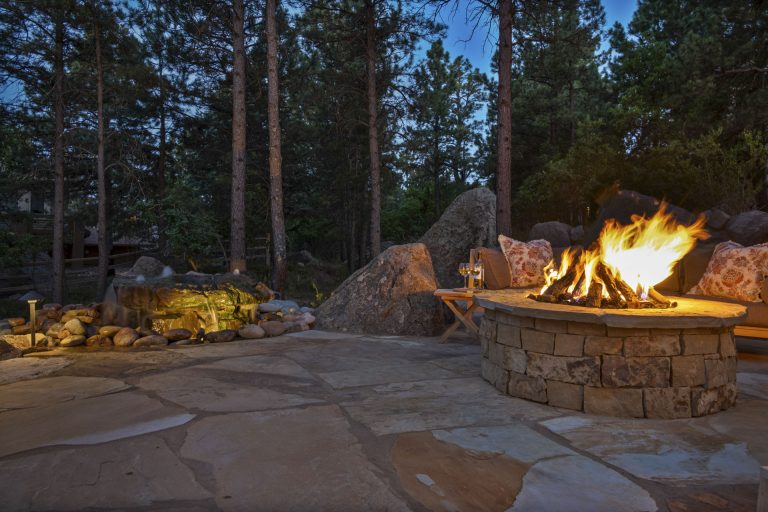 Evening fire pit with stone seating area surrounded by landscaping lights and tall trees