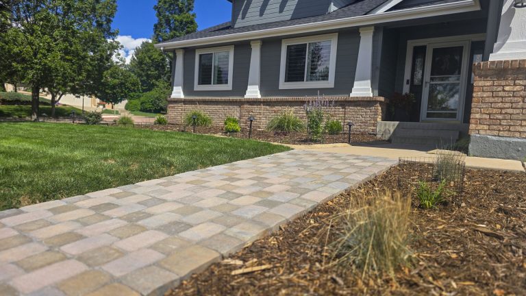 New brick paver walkway leading to blue gray house with landscaping
