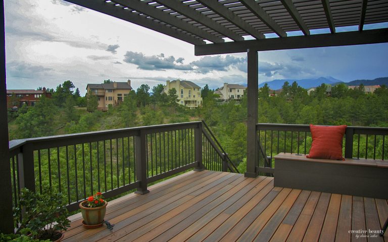 Elevated wooden deck with metal railing under pergola roof overlooking forested valley and distant mountains