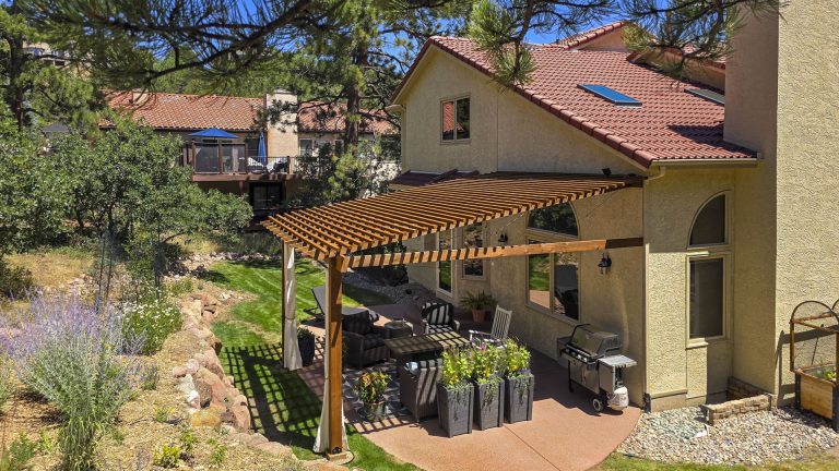 Backyard patio with wooden pergola shade structure attached to tan stucco house with terracotta tile roof
