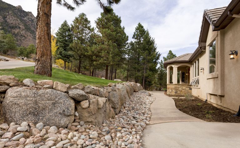 Modern house with stone exterior beside a curved driveway lined with river rocks and mature pine trees
